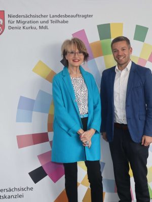 Nina Dieckmann, Vorstandsvorsitzende der Nina.Dieckmann-Stiftung mit Deniz Kurku, Landebeauftragter für Migration und Teilhabe vor Banner des Landesbeauftragten